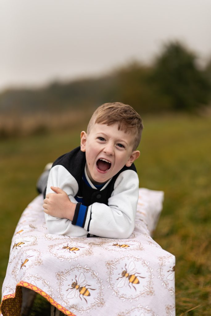 A boy lies on a bench, on a picnic blanket covered in pictures of bees. He is lying on his front, crossed arms and is shouting something towards you. In the background is a blurred meadow with trees.