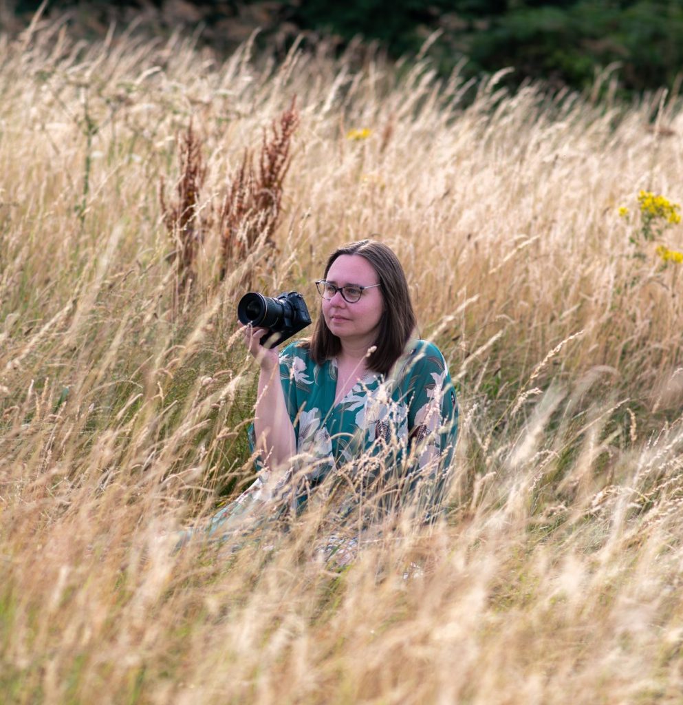 A female photographer wearing glasses and a green flowery dress sitting in a field of long golden grass with her camera in her hand. Both the photographer and the camera are facing your left