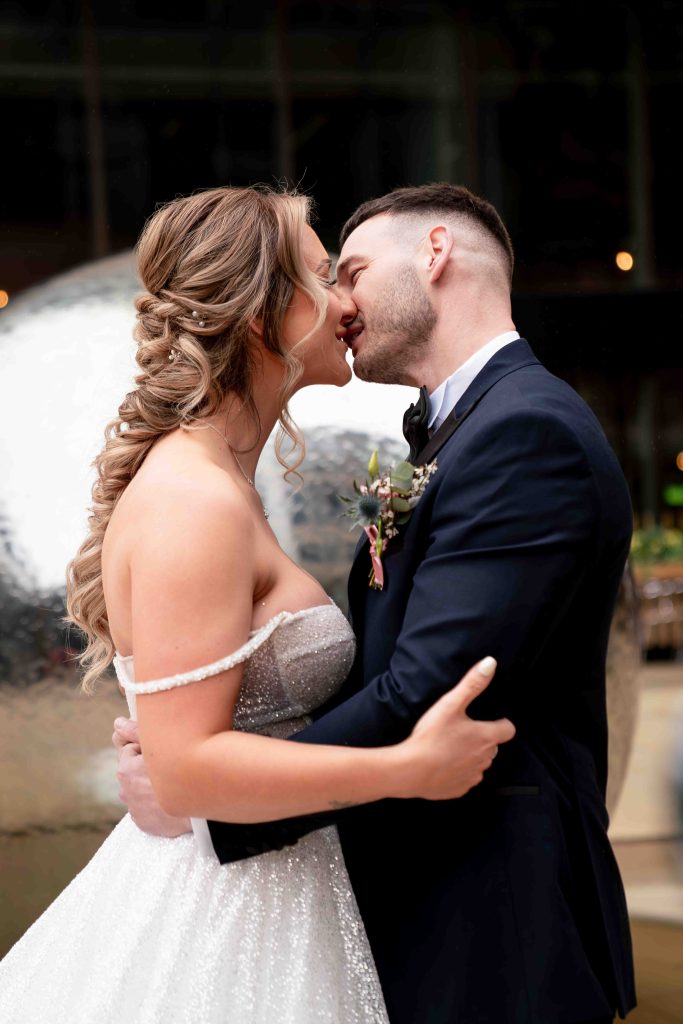 A newly married man and woman kissing. She has a long braid and is wearing a sparkly white dress, with straps half way down her arm. He is wearing a black tuxedo with a wildflower buttonhole. He has his hands on her waist and hers are on his arm. In the background is a blurred metal ball fountain.