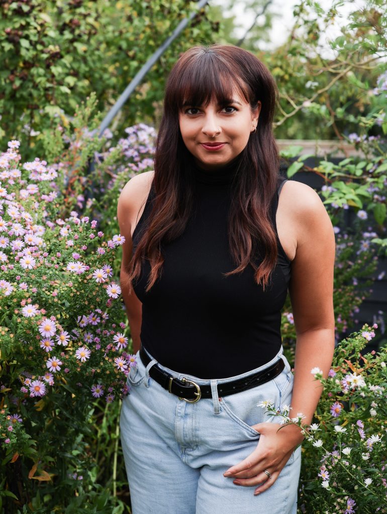 woman wearing a black sleeveless top and pale blue jeans, stands with one hand in her pocket looking towards you. She has long brown dark hair with a fringe and light brown skin. She is standing in front of a green bush with light pink flowers.