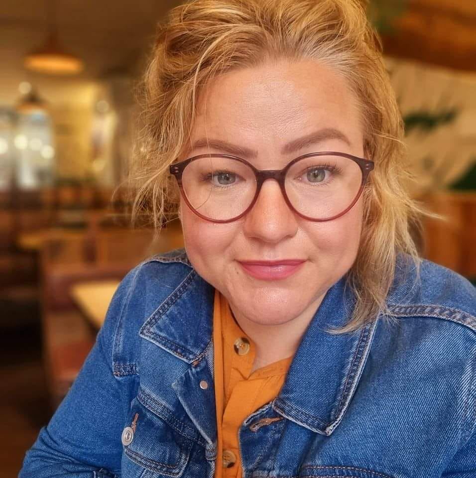 A woman with glasses looking at you. She is wearing a blue denim jacket with an orange top underneath. From the blurred background it is clear she is sitting in a restaurant.