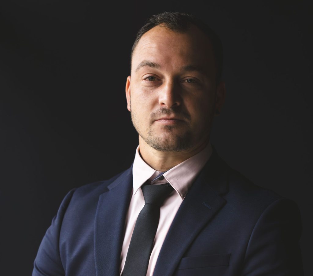 A man in a black suit and tie. He is looking at you. His left side is in darkness, whilst his left in light. He is fastening a cufflink