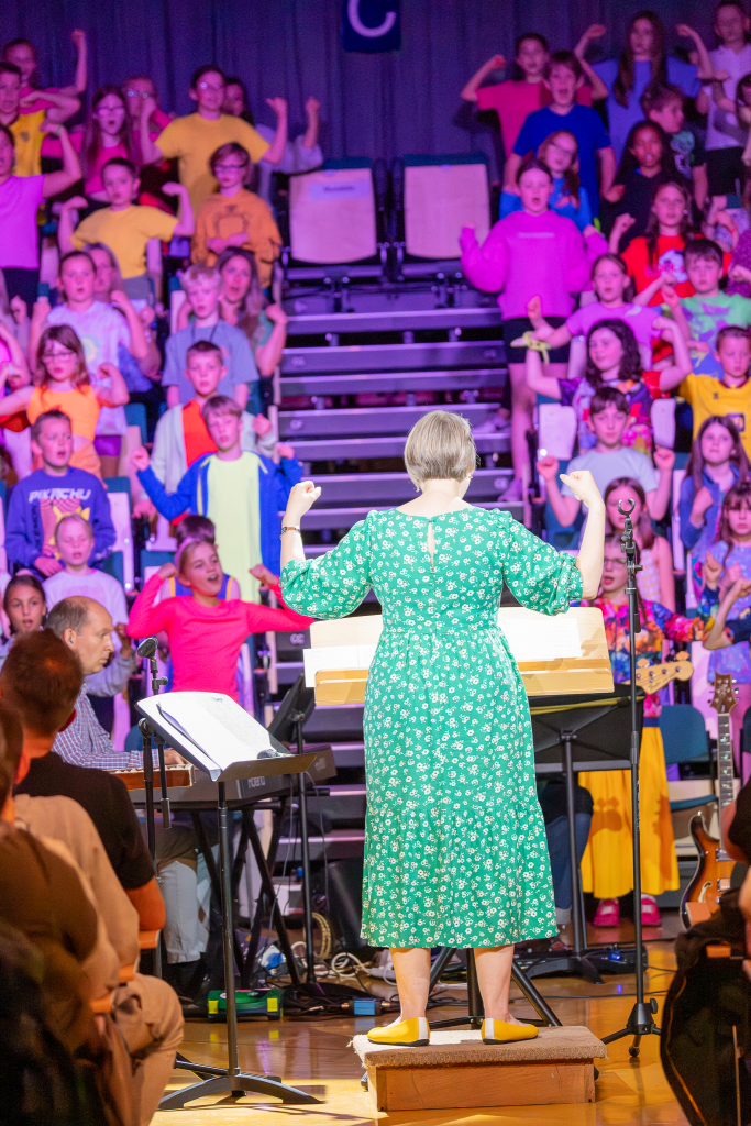 A female conductor in a green dress, facing away from you, conducts child singers wearing brightly coloured clothing. The children are standing in the seat aisles of a large concert theatre, singing. The lighting on the children is purple.